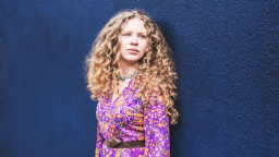 Image of Becca James. She has long curly hair, is wearing a purple, orange and pink floral dress, and is standing against a dark blue wall. 